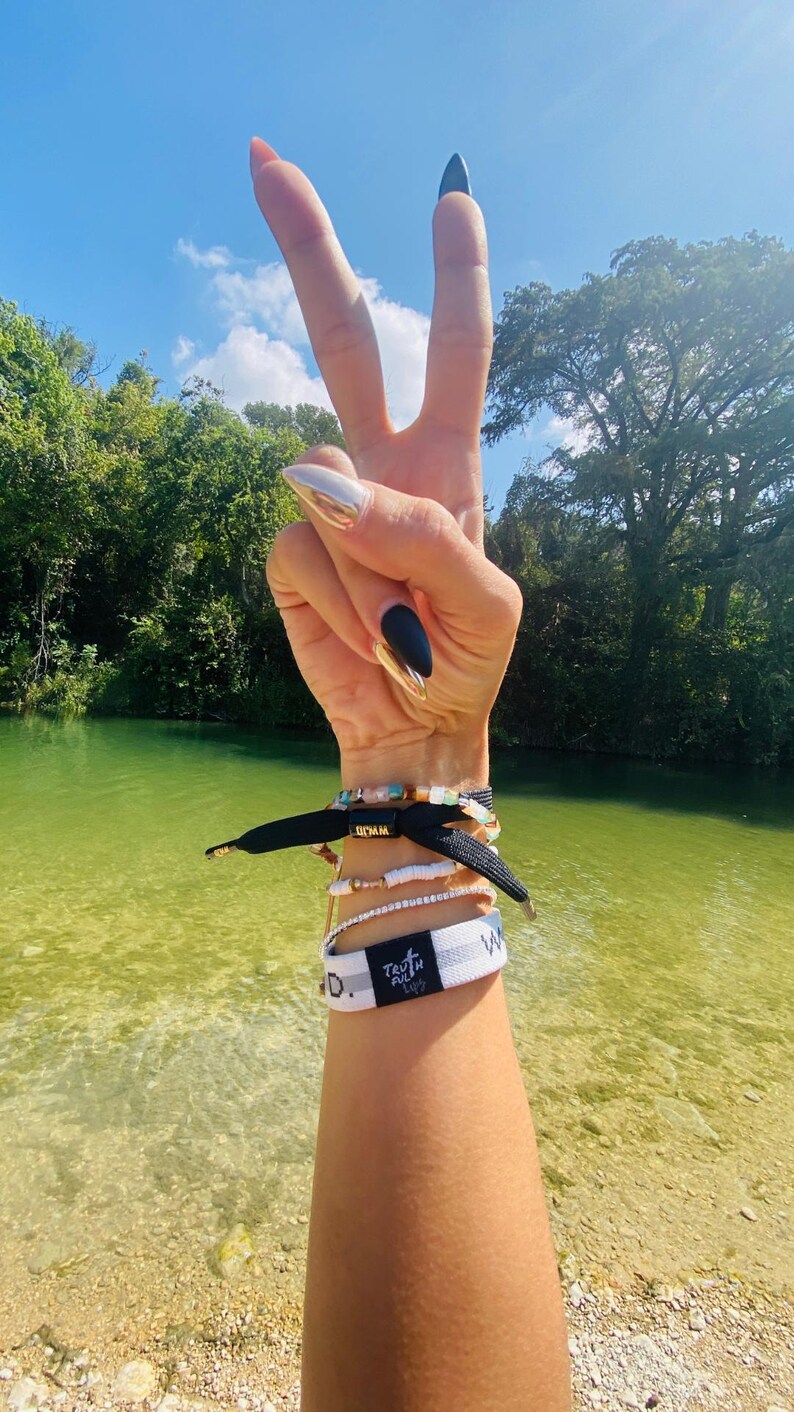May include: A person's arm with black and white bracelets, including one that says "Truthfully" and another that says "W.O.W.". The person is making a peace sign with their hand, and their nails are painted black and silver.
