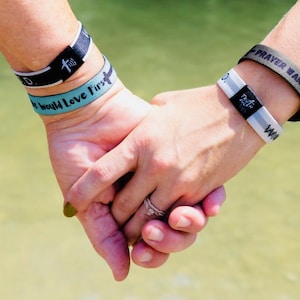 May include: Two people holding hands, each wearing multiple wristbands. The wristbands on the left hand are black with white text that reads "God" and "Family Love First". The wristbands on the right hand are black and white with the text "Testimony" and a green wristband with the text "Prayer Warrior".