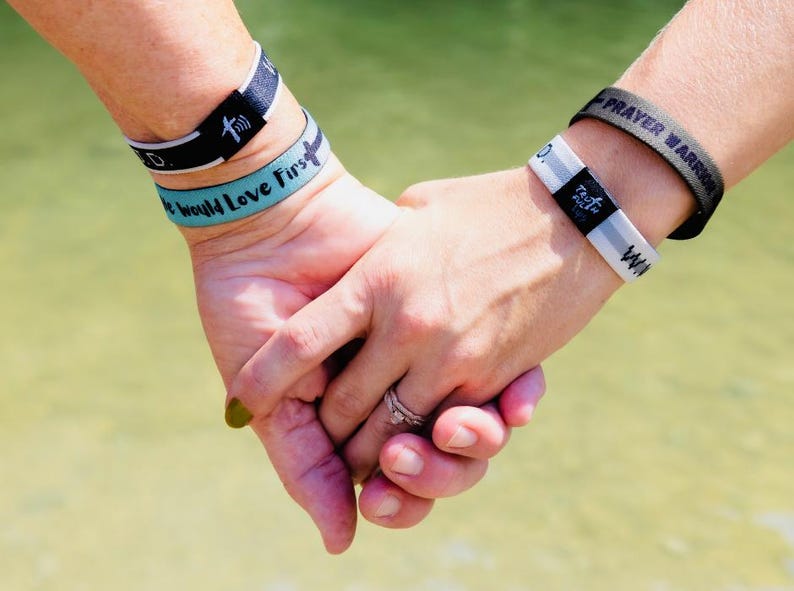May include: Two people holding hands, each wearing multiple wristbands. The wristbands on the left hand are black with white text that reads "God" and "Family Love First". The wristbands on the right hand are black and white with the text "Testimony" and a green wristband with the text "Prayer Warrior".