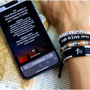 May include: A smartphone displaying a Bible verse on a screen with a sunset background. The verse reads "Cast all your anxiety on him because he cares for you." 1 Peter 5:7 (NIV). The phone is lying on an open book with a feather bookmark. A wrist with several bracelets is visible in the background. The bracelets are black with white lettering and include the words "walk in love" and "EPH 5:2".