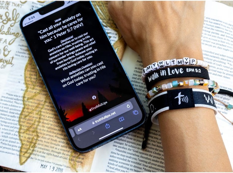 May include: A smartphone displaying a Bible verse on a screen with a sunset background. The verse reads "Cast all your anxiety on him because he cares for you." 1 Peter 5:7 (NIV). The phone is lying on an open book with a feather bookmark. A wrist with several bracelets is visible in the background. The bracelets are black with white lettering and include the words "walk in love" and "EPH 5:2".