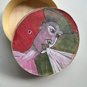 Könnte beinhalten: Eine runde, offene Holzkiste mit Deckel, der eine abstrakte Illustration zeigt. Das Kunstwerk auf dem Deckel zeigt ein Personenprofil in Rosa, Lila und Grün vor rotem Hintergrund. Die Kiste hat eine natürliche Holzfarbe.
