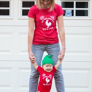 May include: A woman and a child wearing red shirts with a white rooster logo and the words 'Hot Like Fire' and 'Sriracha' in white. The child is wearing a green hat. The woman is wearing blue jeans and the child is wearing a red onesie. The woman and child are standing in front of a white garage door.