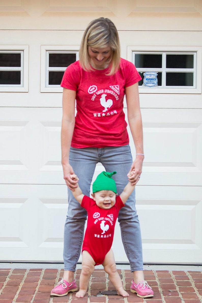 May include: A woman and a child wearing red shirts with a white rooster logo and the words 'Hot Like Fire' and 'Sriracha' in white. The child is wearing a green hat. The woman is wearing blue jeans and the child is wearing a red onesie. The woman and child are standing in front of a white garage door.