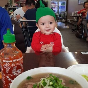 May include: A red and green baby outfit with a green hat. The baby is sitting in a high chair at a table with a bowl of pho and a bottle of sriracha hot chili sauce.