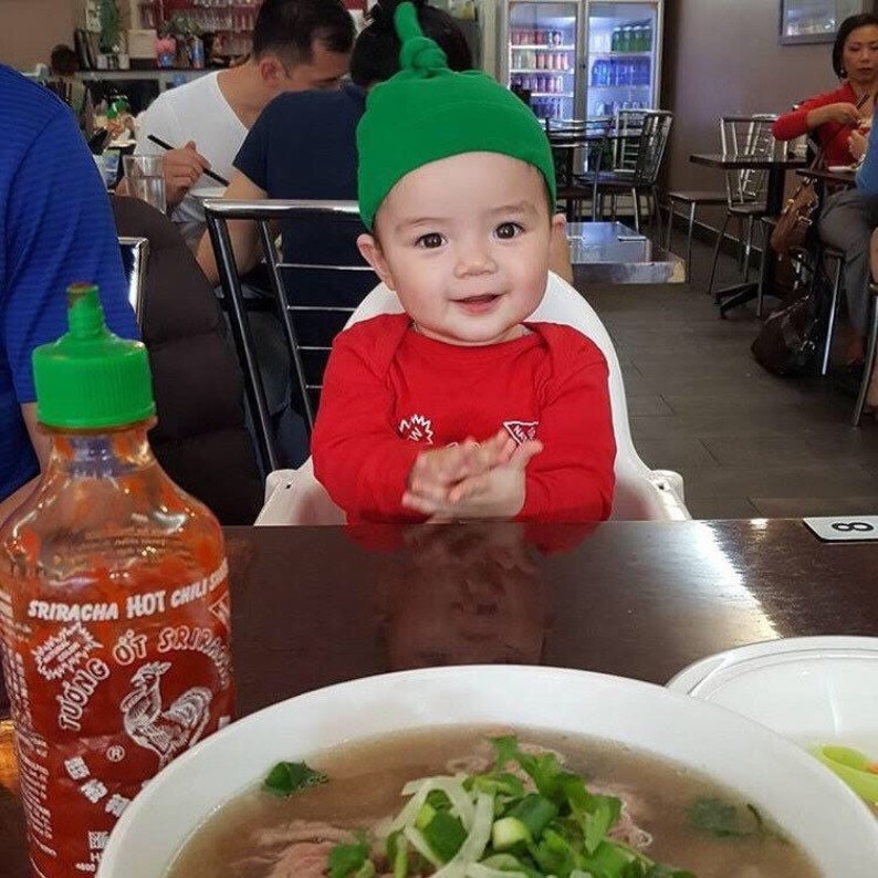 May include: A red and green baby outfit with a green hat. The baby is sitting in a high chair at a table with a bowl of pho and a bottle of sriracha hot chili sauce.