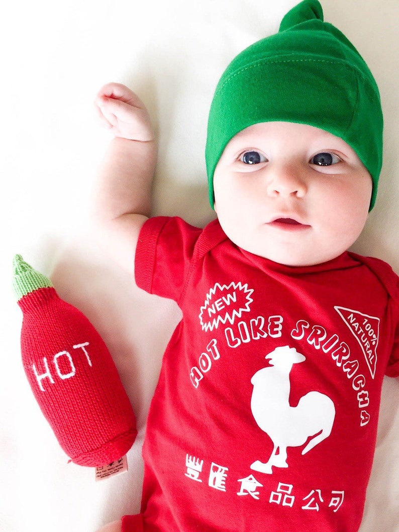May include: A baby wearing a red shirt with a white rooster and the words 'Hot Like Sriracha' and '100% Natural' printed on it. The baby is wearing a green beanie and holding a red knitted hot sauce bottle that says 'Hot'.