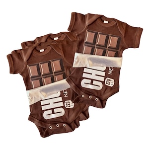 May include: Two brown short-sleeve baby bodysuits featuring a graphic of a chocolate bar. The design includes a silver foil wrapper detail, the text "CHO" in large white letters, and a small "220 cal" label on the lower front section.