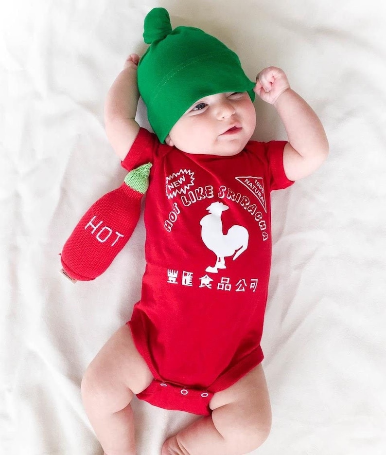 May include: A baby wearing a red bodysuit with a white rooster and the text 'Hot Like Sriracha' and 'New Natural 100%  Sriracha' in a white font. The baby is also wearing a green hat and holding a red plush pepper with the word 'Hot' on it.
