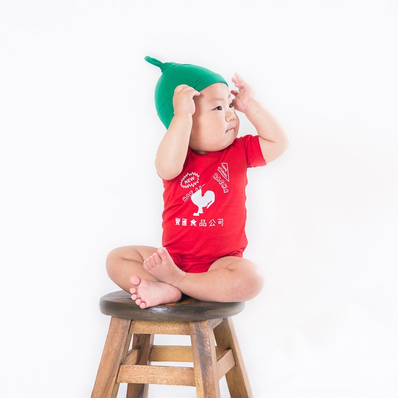 May include: A baby wearing a red bodysuit with a white rooster logo and the words 'NEW! HOT SAUCE' and '닭갈비 食品公司' in white. The baby is also wearing a green hat and is sitting on a wooden stool.