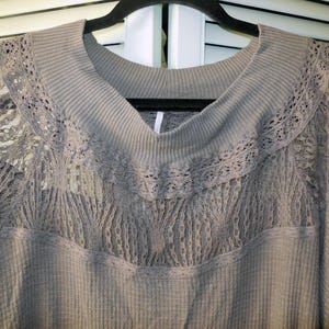 May include: A gray long-sleeved top with a scoop neck and lace detailing. The top features a waffle knit texture and delicate lace inserts across the chest and shoulders. The top is hanging on a black hanger.