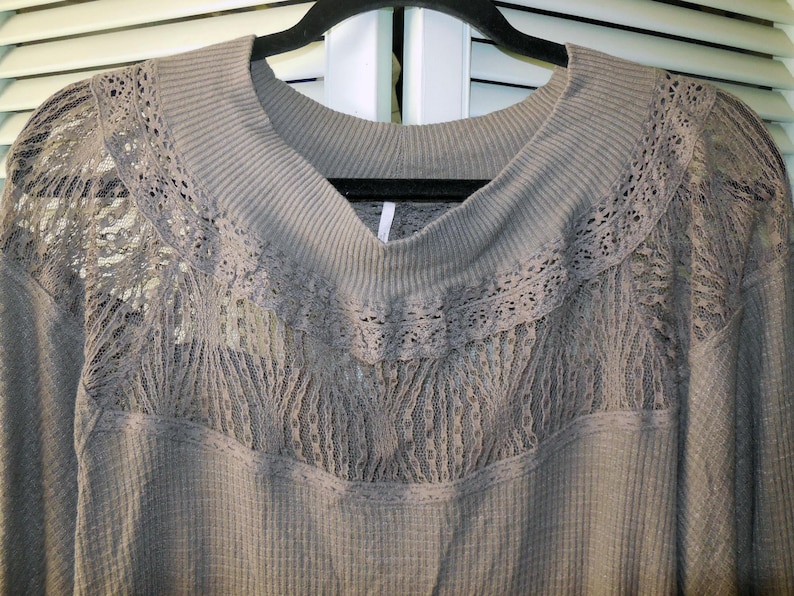 May include: A gray long-sleeved top with a scoop neck and lace detailing. The top features a waffle knit texture and delicate lace inserts across the chest and shoulders. The top is hanging on a black hanger.