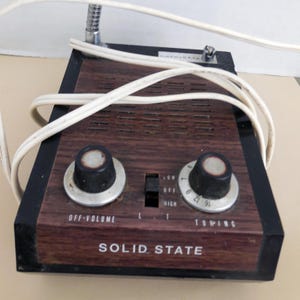 Puede incluir: Radio de estado sólido vintage con un panel frontal de veta de madera y borde negro. Cuenta con dos perillas "OFF-VOLUME" y "TUNING", un interruptor para frecuencias "LOW" y "HIGH" y el texto "SOLID STATE". También se ve un cable blanco y una antena.