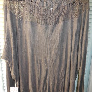 May include: A long-sleeved, taupe-colored top with a waffle knit texture. The top features a high neckline and a delicate lace panel across the upper back. The sleeves are long with ribbed cuffs. A tag is attached to the garment.