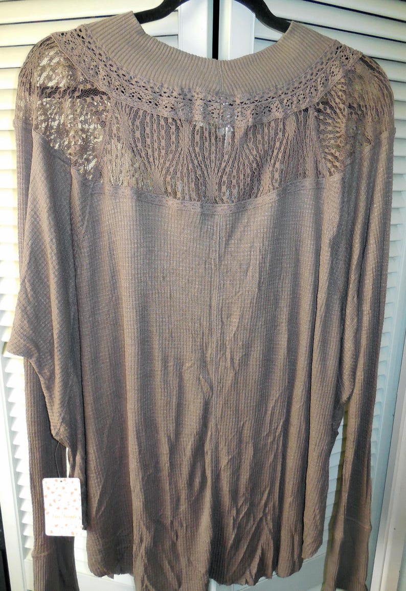 May include: A long-sleeved, taupe-colored top with a waffle knit texture. The top features a high neckline and a delicate lace panel across the upper back. The sleeves are long with ribbed cuffs. A tag is attached to the garment.