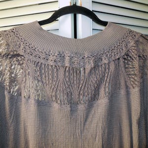 May include: A taupe-colored long-sleeved top with a ribbed texture. The top features a high neckline and a delicate lace panel across the upper back and shoulders, adding a touch of elegance to the design.