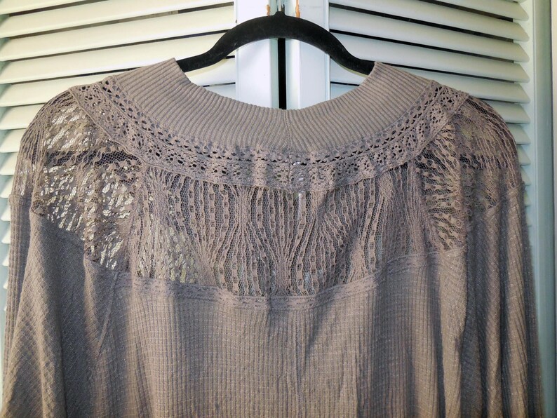 May include: A taupe-colored long-sleeved top with a ribbed texture. The top features a high neckline and a delicate lace panel across the upper back and shoulders, adding a touch of elegance to the design.