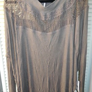 May include: A long-sleeved, taupe-colored shirt with a waffle-knit texture. The shirt features a V-neck and a decorative lace panel across the chest and shoulders. The sleeves have a fitted cuff. The shirt is hanging on a black hanger.