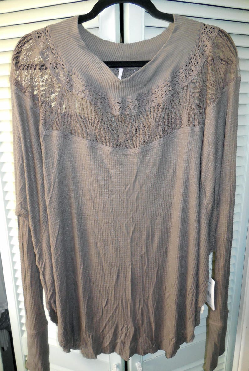 May include: A long-sleeved, taupe-colored shirt with a waffle-knit texture. The shirt features a V-neck and a decorative lace panel across the chest and shoulders. The sleeves have a fitted cuff. The shirt is hanging on a black hanger.