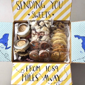 May include: A cardboard box with a yellow and white striped pattern. The box is open and contains various baked goods, including cookies, brownies, and a jar of homemade jam. The box is decorated with a handwritten message that says "Sending you sweets from 1089 miles away".