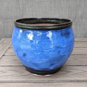 May include: A round, blue ceramic planter with a dark rim and base. The planter has a textured, glazed finish with shades of blue and black. The interior is a lighter brown color. The planter is sitting on a wooden surface.