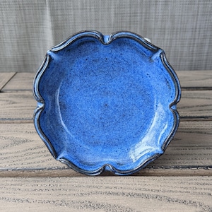 May include: A handcrafted, blue ceramic dish with a unique scalloped edge. The dish has a speckled texture and a dark rim, set against a rustic wooden surface. This decorative piece is ideal for serving or display.