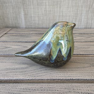May include: A ceramic bird figurine with a glossy finish. The bird has a rounded body, a pointed beak, and a small tail. The colors include shades of green, blue, and brown. The figurine is placed on a wooden surface.