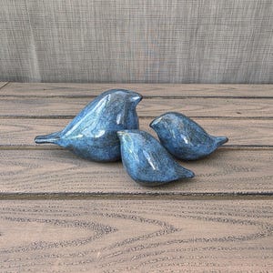 May include: Three blue ceramic bird figurines of varying sizes. The birds have a glossy finish and are arranged on a wooden surface. The color is a mottled blue with darker accents, giving them a textured appearance. The birds are a decorative item.