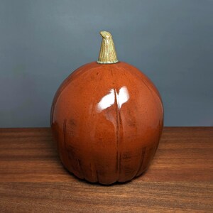 May include: A ceramic pumpkin with a glossy, reddish-brown finish. The pumpkin has vertical grooves and a textured, light green and tan stem. The decorative gourd sits on a wooden surface against a blue-gray background.
