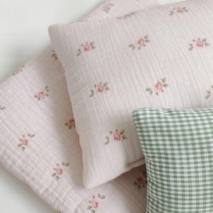 Könnte beinhalten: Ein hellrosa rechteckiges Kissen und eine passende gefaltete Decke, beide mit einem kleinen, sich wiederholenden Blumenmuster aus rosa Rosen und grünen Blättern. Ein kleineres rechteckiges Kissen mit grün-weißem Vichy-Karo liegt davor.