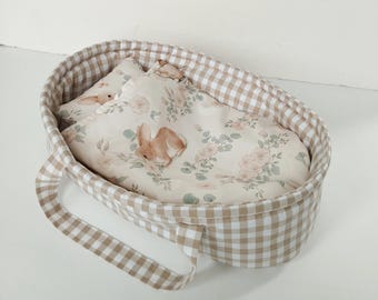 Puede incluir: Una cuna de muñeca de gingham beige y blanco con un forro de estampado floral y un conejito de peluche dentro.