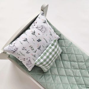 Op de afbeelding: Een wit poppenbed met een groene, gestikte matras en twee kussens. Een kussen heeft een witte achtergrond met een bloemenpatroon en konijnen, het andere kussen is groen-wit geruit.