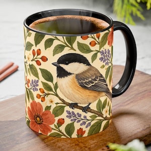 May include: A ceramic mug with a black handle and interior, featuring a cream-colored exterior decorated with a pattern of a black-capped chickadee perched on a branch surrounded by orange flowers, green leaves, and small red berries.