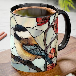 May include: A black-handled coffee mug featuring a stained-glass style design of a bird perched on a branch with red flowers. The bird has a black head, white cheeks, and brown and yellow body. The mug is filled with coffee.