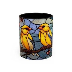 Puede incluir: Una taza de caf&eacute; de cer&aacute;mica con un dise&ntilde;o de vidriera que presenta dos p&aacute;jaros amarillos posados en una rama marr&oacute;n. Los p&aacute;jaros tienen picos naranjas y est&aacute;n sobre un fondo azul y gris. El interior de la taza es negro.