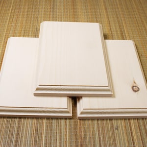 May include: Four unfinished wooden plaques with decorative edges. The rectangular plaques are made of light-colored wood and are arranged on a textured, light brown surface. The plaques have a raised, detailed border.