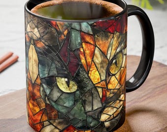 May include: Black ceramic mug featuring a stained-glass cat face design in shades of orange, brown, green, and yellow. The mug has a black handle and is filled with a dark liquid. The mug is sitting on a wooden surface.