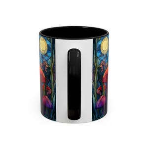 Peut inclure: Une tasse &agrave; caf&eacute; en c&eacute;ramique avec un int&eacute;rieur et une anse noirs, et un ext&eacute;rieur blanc. La tasse pr&eacute;sente une illustration color&eacute;e de champignons, de plantes et d'une lune. Le design est dans les tons de rouge, violet, bleu et jaune.