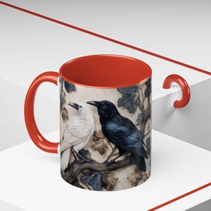 May include: A ceramic mug with a red interior and handle. The exterior features a detailed relief of two ravens, one white and one black, perched on a branch with leaves. The mug is set against a white and red background.