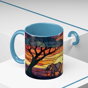 Puede incluir: Taza de cer&aacute;mica con asa e interior azules. El exterior presenta un dise&ntilde;o de vidriera de una escena de sabana africana con un &aacute;rbol, un elefante y una puesta de sol en tonos naranja, amarillo y morado.