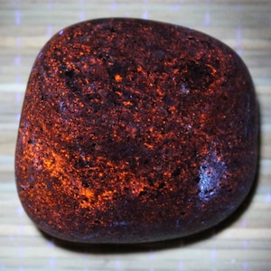 May include: A rounded, dark brown stone with vibrant orange and blue speckles. The stone has a smooth, polished appearance, with the orange specks appearing to glow against the darker background. The overall shape is irregular, with rounded edges.