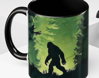 May include: Black ceramic mug featuring a Bigfoot silhouette walking through a green forest scene. The mug has a black handle and interior, with a detailed illustration of trees and the legendary creature.