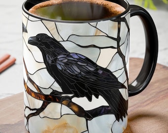 May include: A ceramic mug with a black handle and rim, featuring a stained-glass design of a black raven perched on a branch. The background is a mosaic of white, cream, and gray shapes. The mug is filled with a dark liquid.