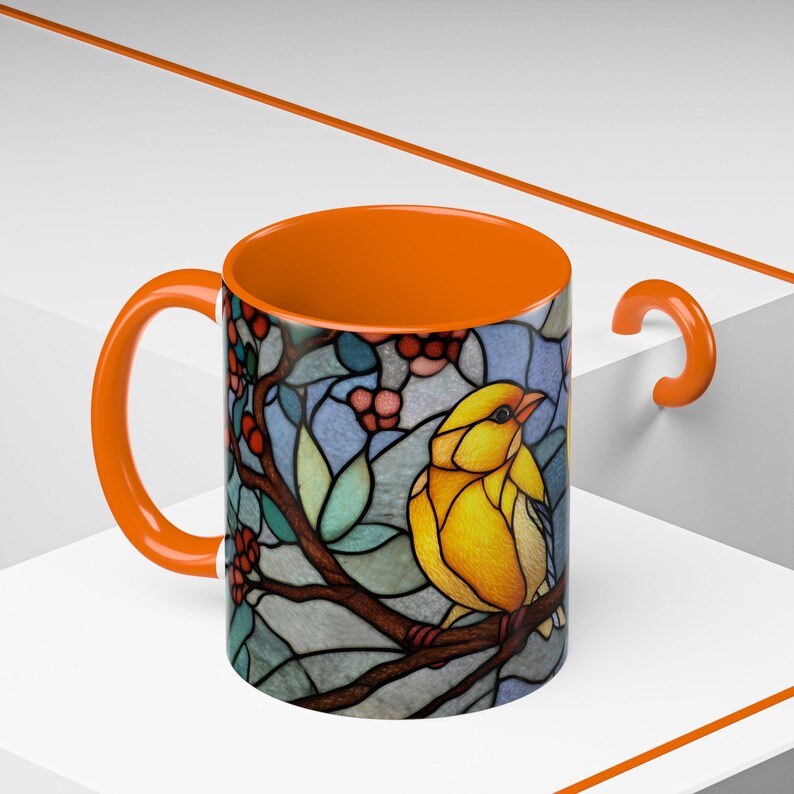 Puede incluir: Taza de cer&aacute;mica naranja con un dise&ntilde;o de vidriera que presenta un p&aacute;jaro amarillo posado en una rama con bayas rojas y hojas verdes. El interior y el asa de la taza tambi&eacute;n son naranjas.