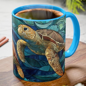 May include: A ceramic coffee mug with a blue handle and rim, featuring a detailed illustration of a sea turtle swimming in blue and green water. The turtle has brown and tan markings. The mug is filled with a dark liquid.
