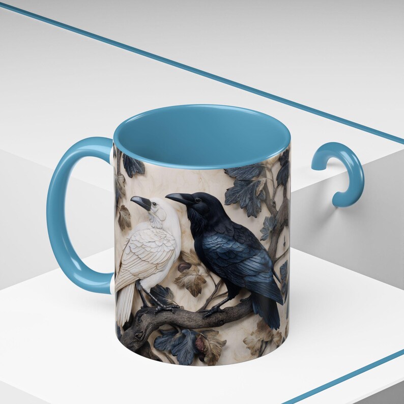 May include: A ceramic mug with a light blue interior and handle. The exterior features a detailed relief of two birds, one white and one black, perched on a branch with leaves. The background has a beige tone.