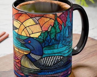 May include: A black-handled coffee mug with a colorful stained-glass design featuring a loon on a lake with a sunset background. The mug is filled with a dark liquid, and sits on a wooden surface.