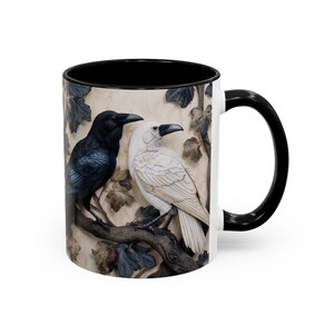 May include: White ceramic mug with a black interior and handle. The mug features a detailed relief of two ravens perched on a branch with leaves. One raven is black, the other is white, both with black beaks.