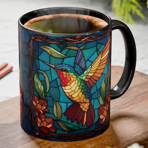 May include: A black coffee mug featuring a colorful stained-glass design of a hummingbird with outstretched wings. The bird is depicted in vibrant hues of red, orange, yellow, green, and blue, set against a teal background. The mug has a black handle and rim.