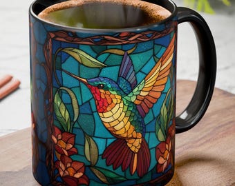 May include: A black coffee mug featuring a colorful stained-glass design of a hummingbird with outstretched wings. The bird is depicted in vibrant hues of red, orange, yellow, green, and blue, set against a teal background. The mug has a black handle and rim.